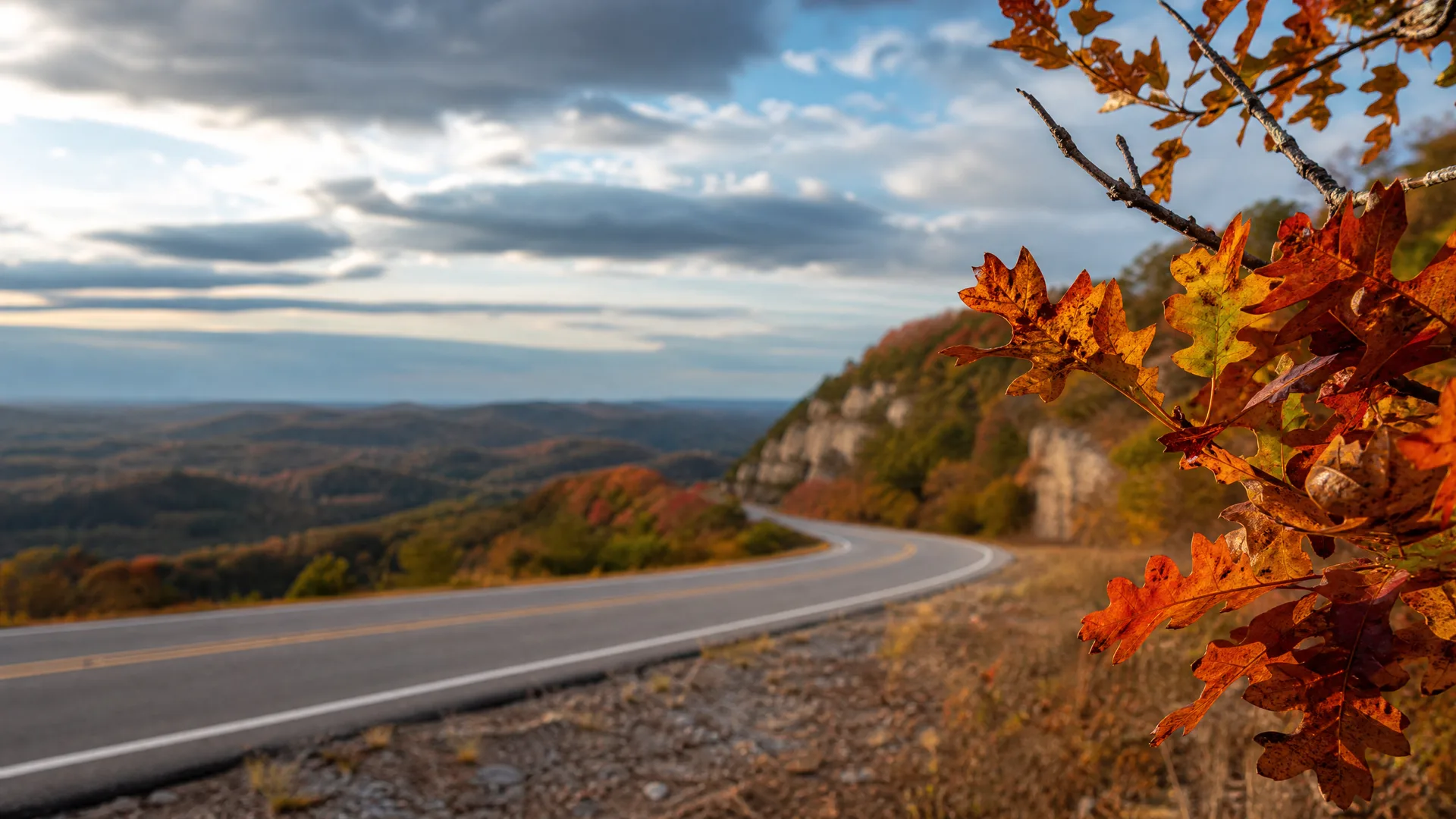 A two-lane paved road curves along a ridge above forested rolling hills, with orange and red oak leaves on a branch in the right foreground and a partly cloudy sky overhead.